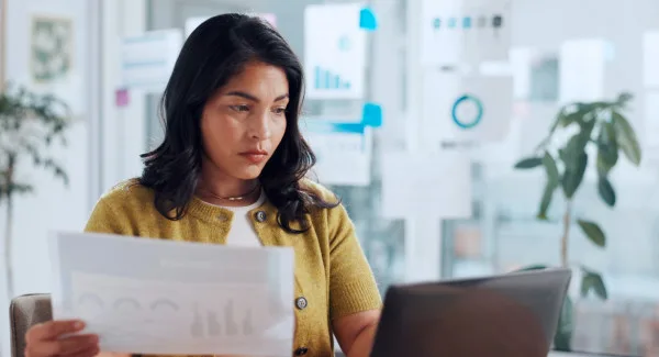 Image of a woman looking at her computer with a piece of paper in her hand